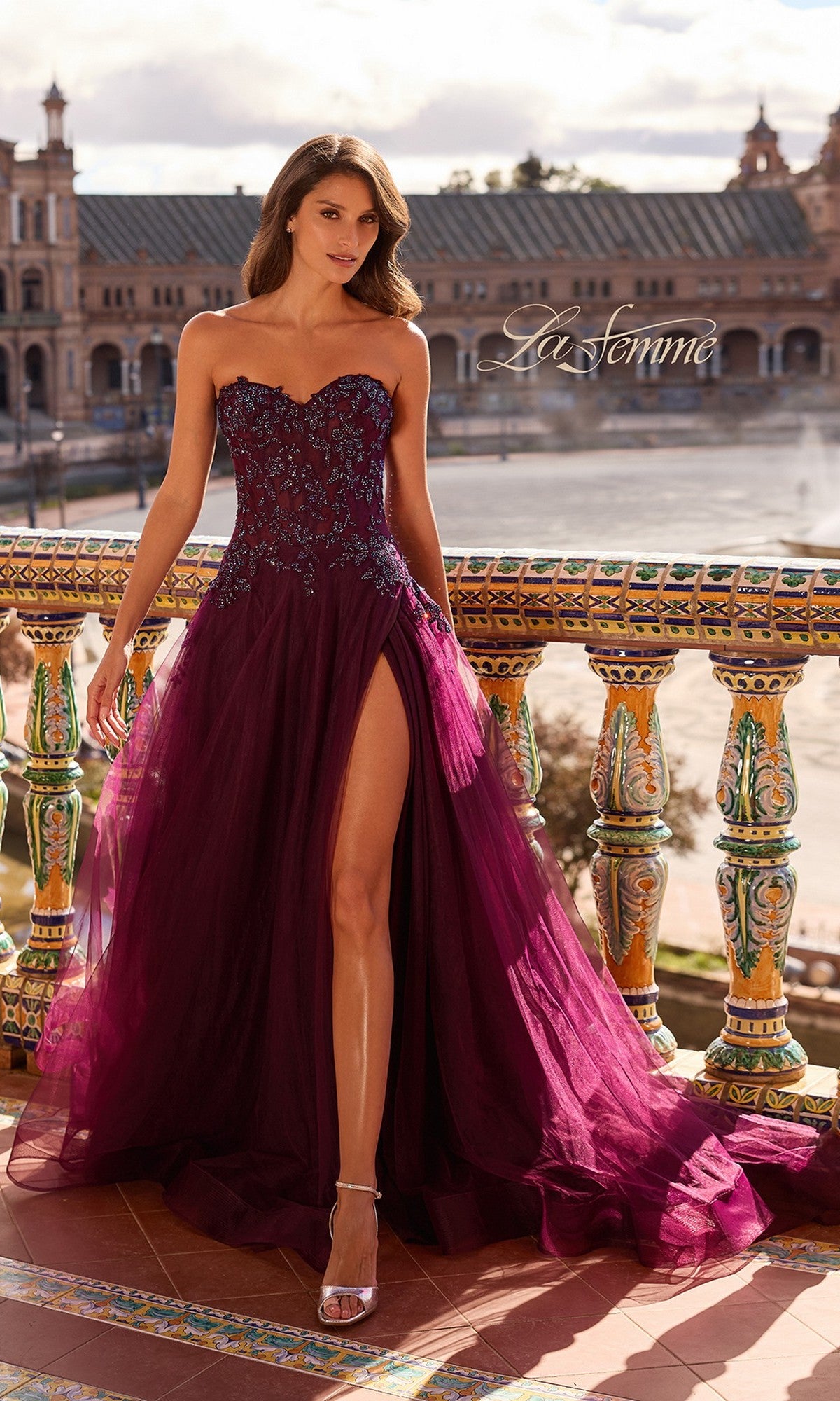 A woman in the Long Prom Dress: La Femme 33551 in Dark Berry, with a strapless beaded lace applique and sheer corset bodice, stands on an ornate balcony against stunning architectural buildings.